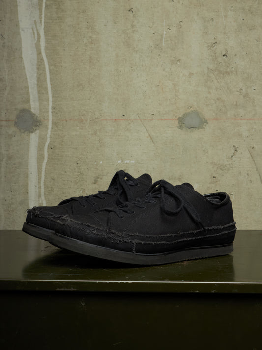 A pair of black low-top lace-up sneakers with a distressed, raw-edge aesthetic, resting on a dark green surface against a textured concrete wall.