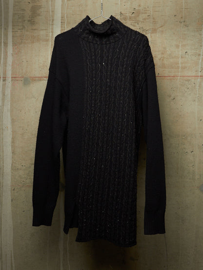 A black knit turtleneck sweater with a textured cable panel down the front, hanging against a textured concrete wall.