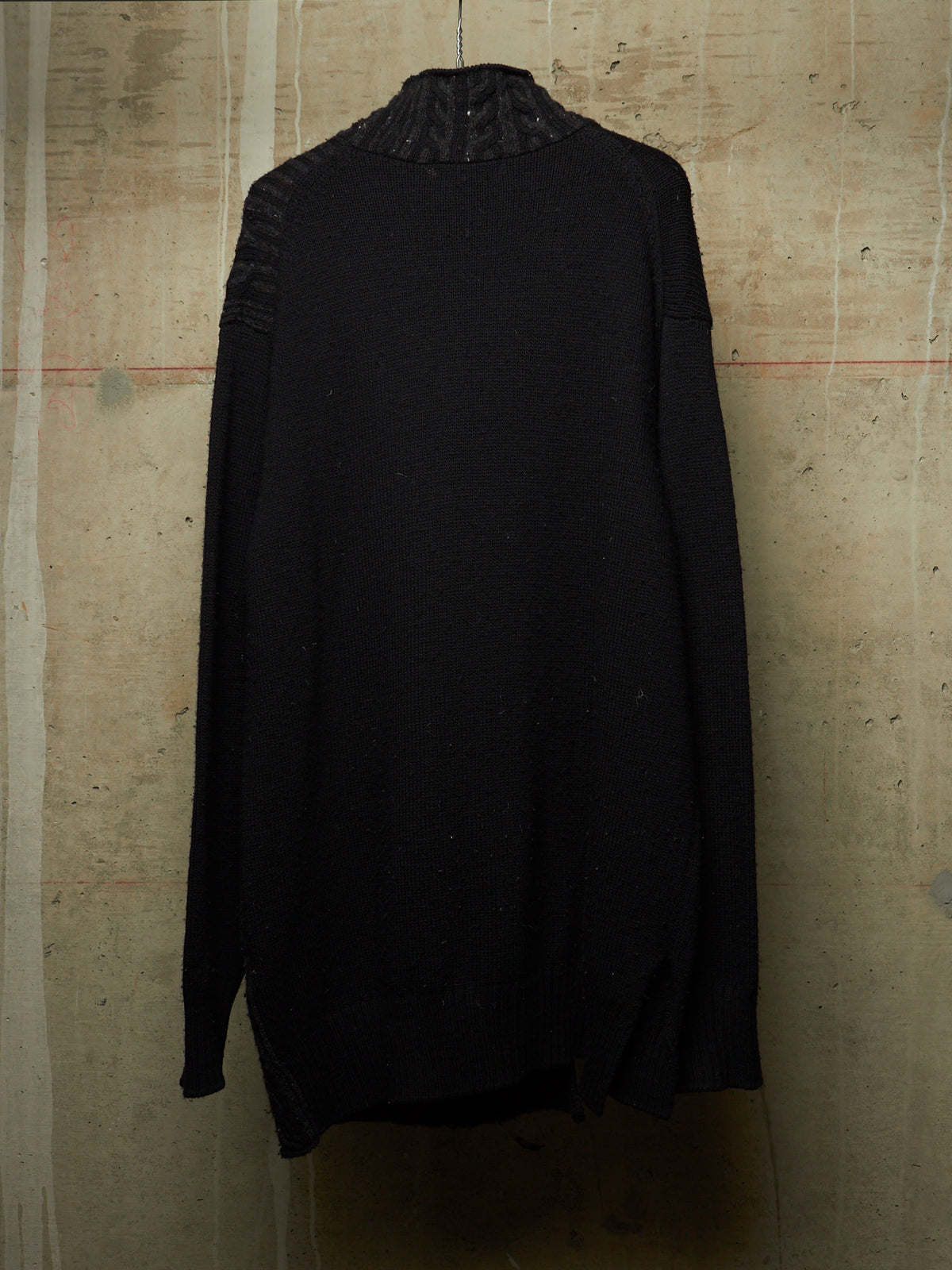 A black knit sweater with a textured, ribbed mock neck and ribbed sleeve cuffs, hanging against a textured concrete wall.