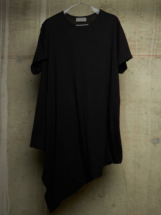 A black short-sleeve t-shirt dress with an asymmetrical hem, hanging on a white hanger against a textured concrete wall.