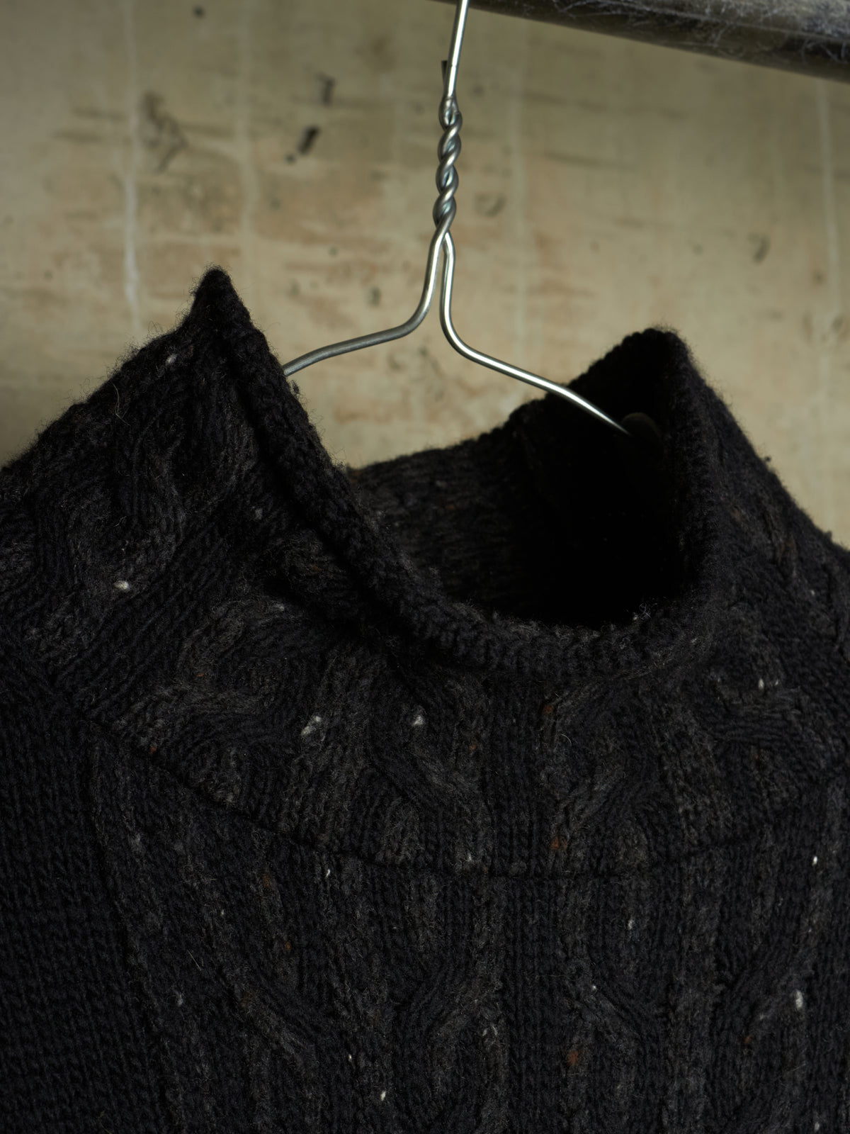 A close-up of a black cable-knit sweater hanging on a metal hanger against a textured beige wall.