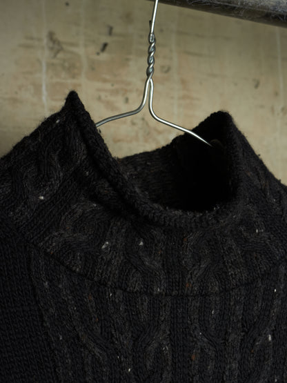 A close-up of a black cable-knit sweater hanging on a metal hanger against a textured beige wall.