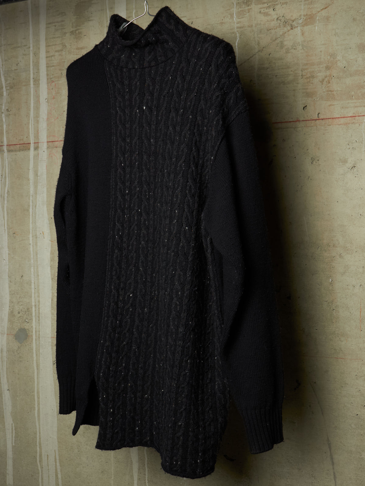 A black long-sleeve knit sweater with a textured cable-knit front panel and solid black sleeves, hanging against a textured concrete wall.