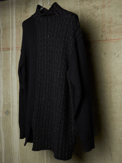 A black long-sleeve knit sweater with a textured cable-knit front panel and solid black sleeves, hanging against a textured concrete wall.
