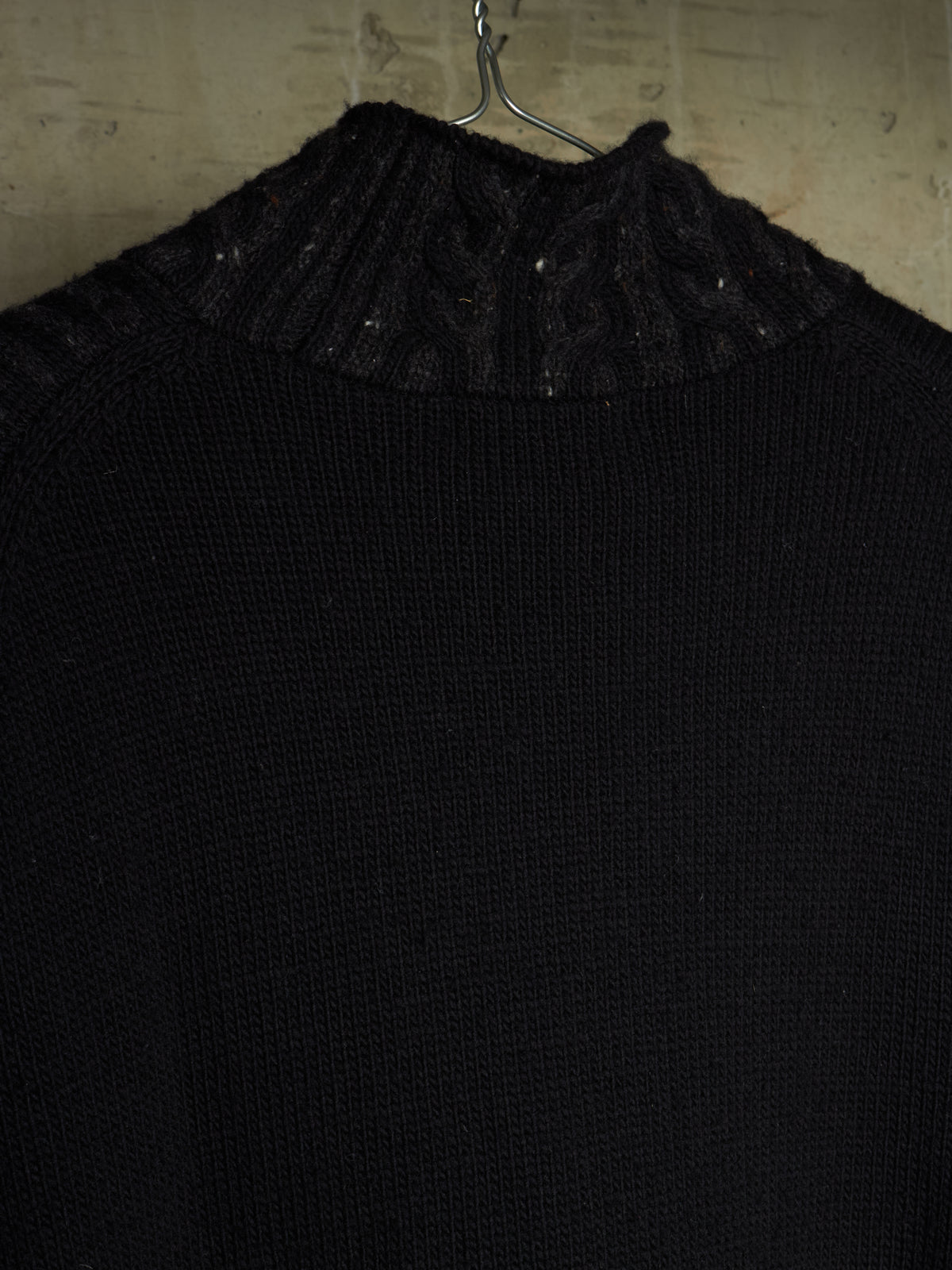 A close-up of a black knit sweater with a textured cable and ribbed mock neck collar, hanging on a metal hanger against a rustic, light-colored wooden wall.