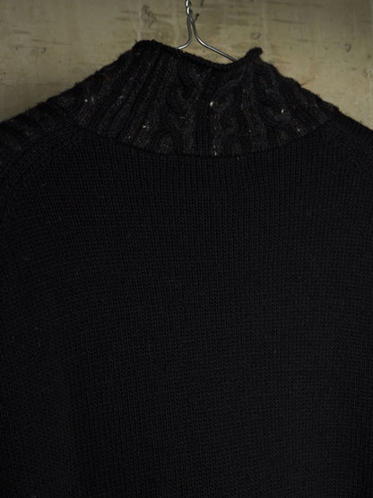 A close-up of a black knit sweater with a textured cable and ribbed mock neck collar, hanging on a metal hanger against a rustic, light-colored wooden wall.