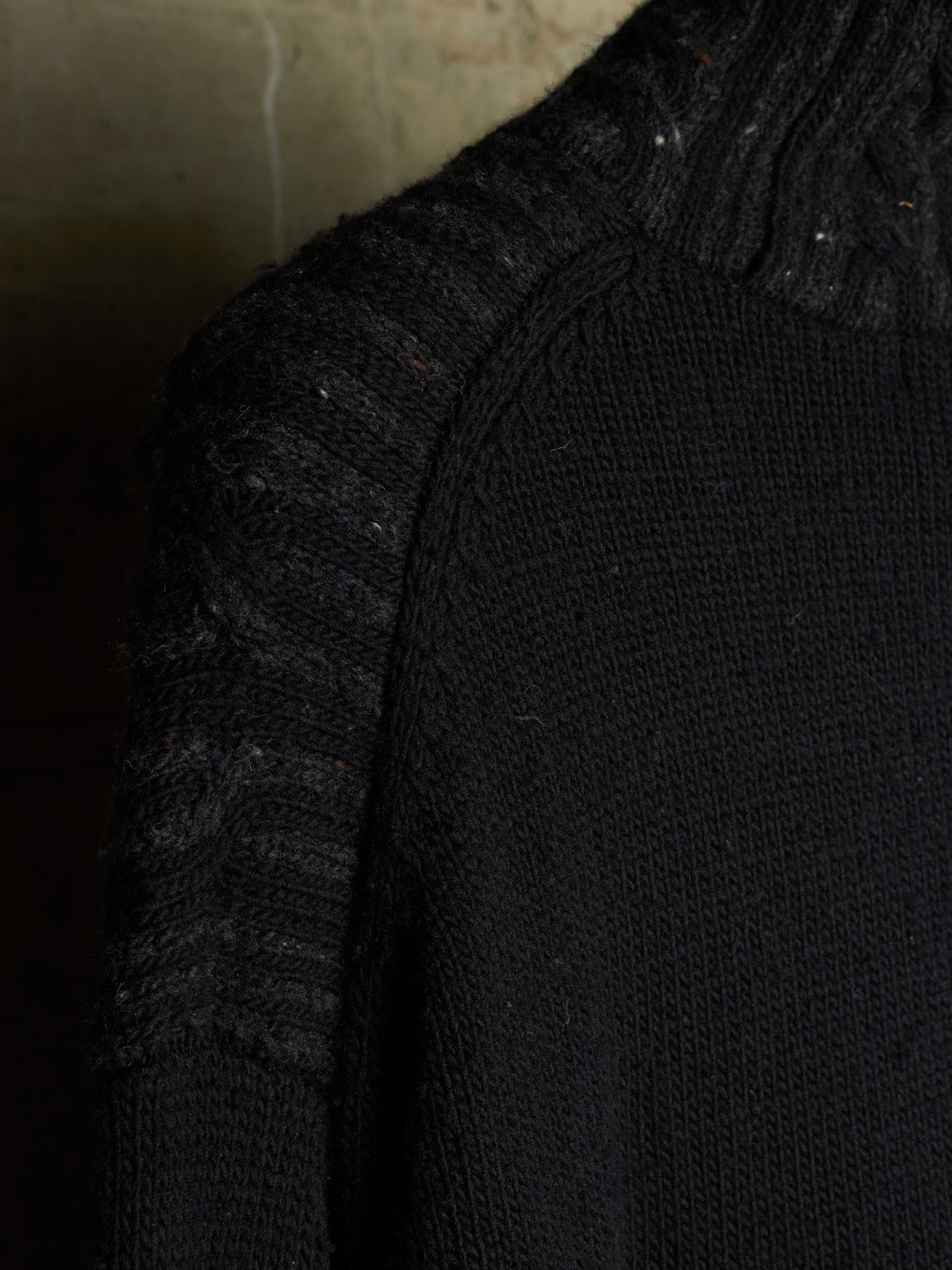 Close-up of a black knit sweater showing the shoulder and ribbed turtleneck area against a dark textured background.