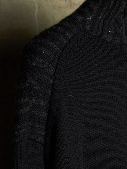 Close-up of a black knit sweater showing the shoulder and ribbed turtleneck area against a dark textured background.