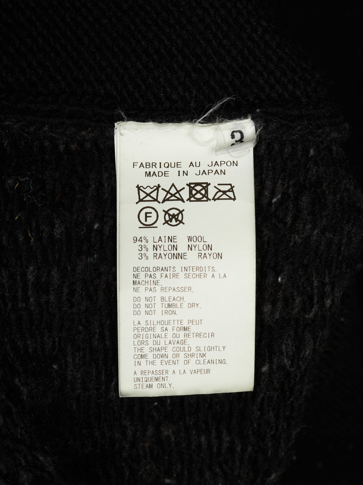A white paper garment care label with laundry care symbols and text, including 'MADE IN JAPAN' and wool/nylon composition details, photographed against a black textured fabric background.