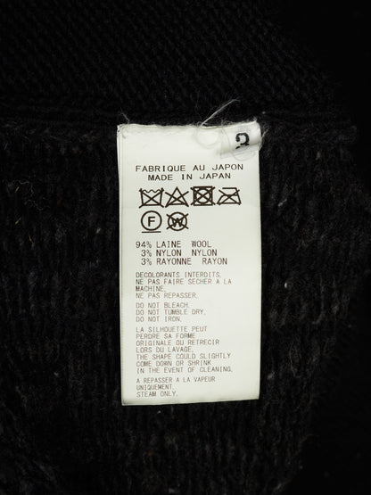 A white paper garment care label with laundry care symbols and text, including 'MADE IN JAPAN' and wool/nylon composition details, photographed against a black textured fabric background.
