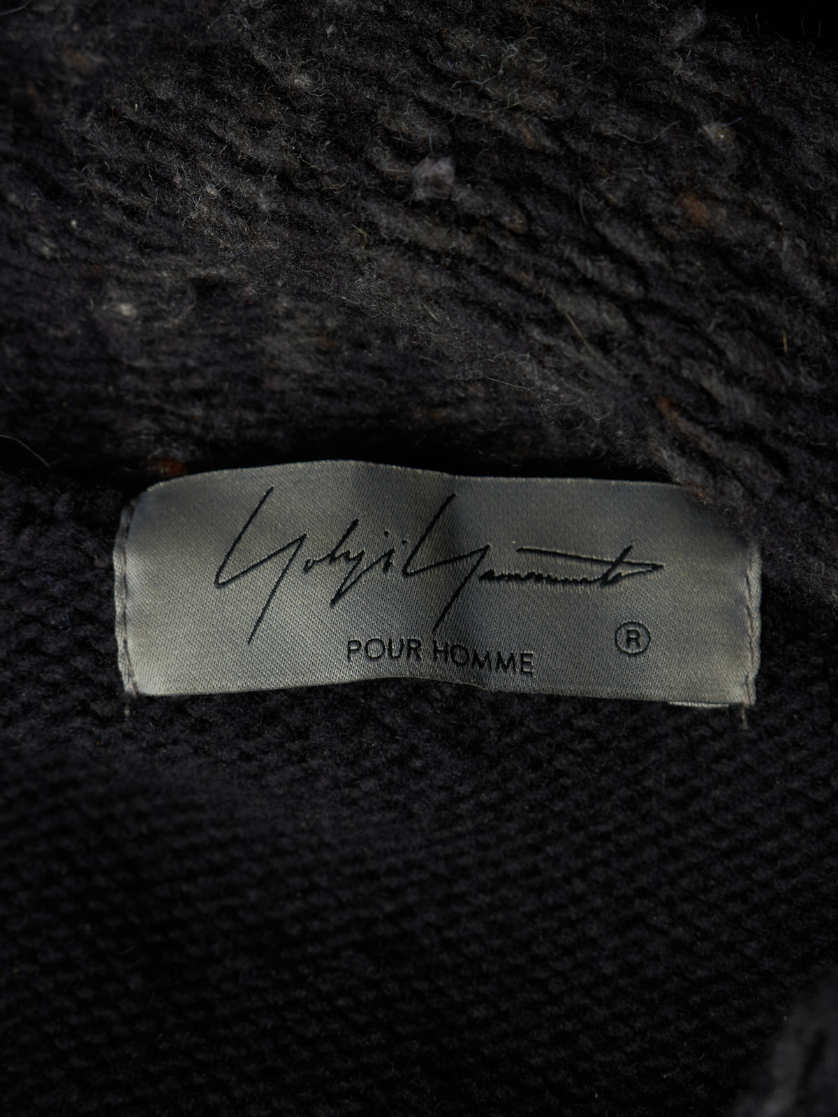 Close-up of a black knit garment label reading 'Ghbirri Ghbirri POUR HOMME' on a dark textured fabric background.