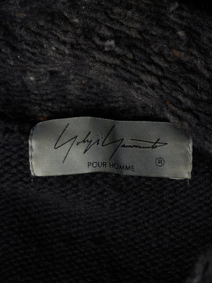 Close-up of a black knit garment label reading 'Ghbirri Ghbirri POUR HOMME' on a dark textured fabric background.