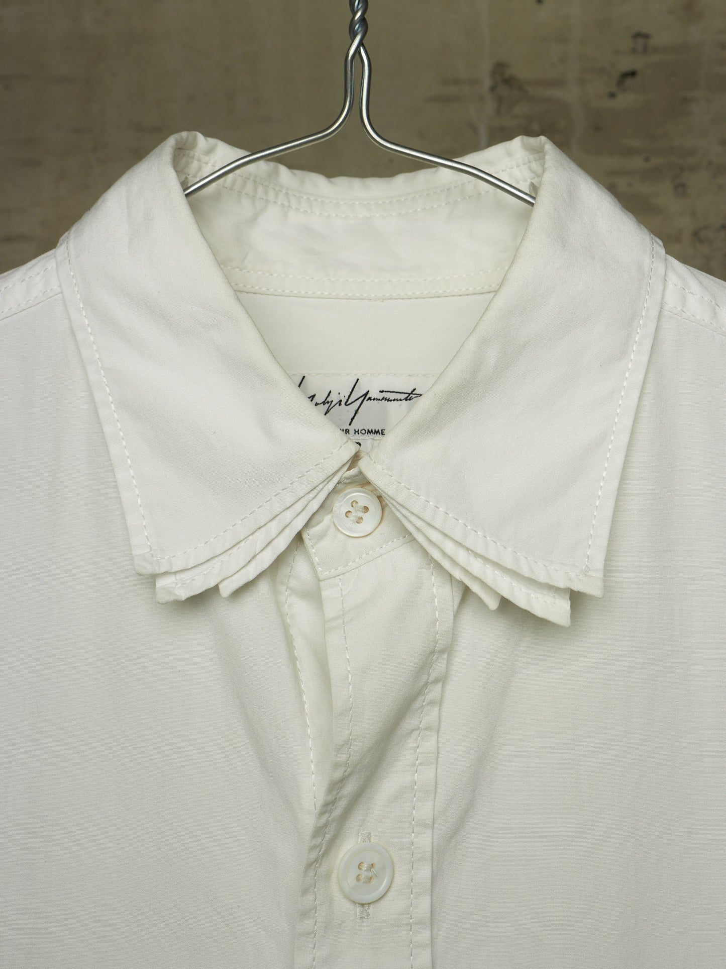 A close-up of a white button-up shirt with a wide, structured collar and decorative pleated detailing at the collar, hanging on a metal hanger against a textured neutral background.