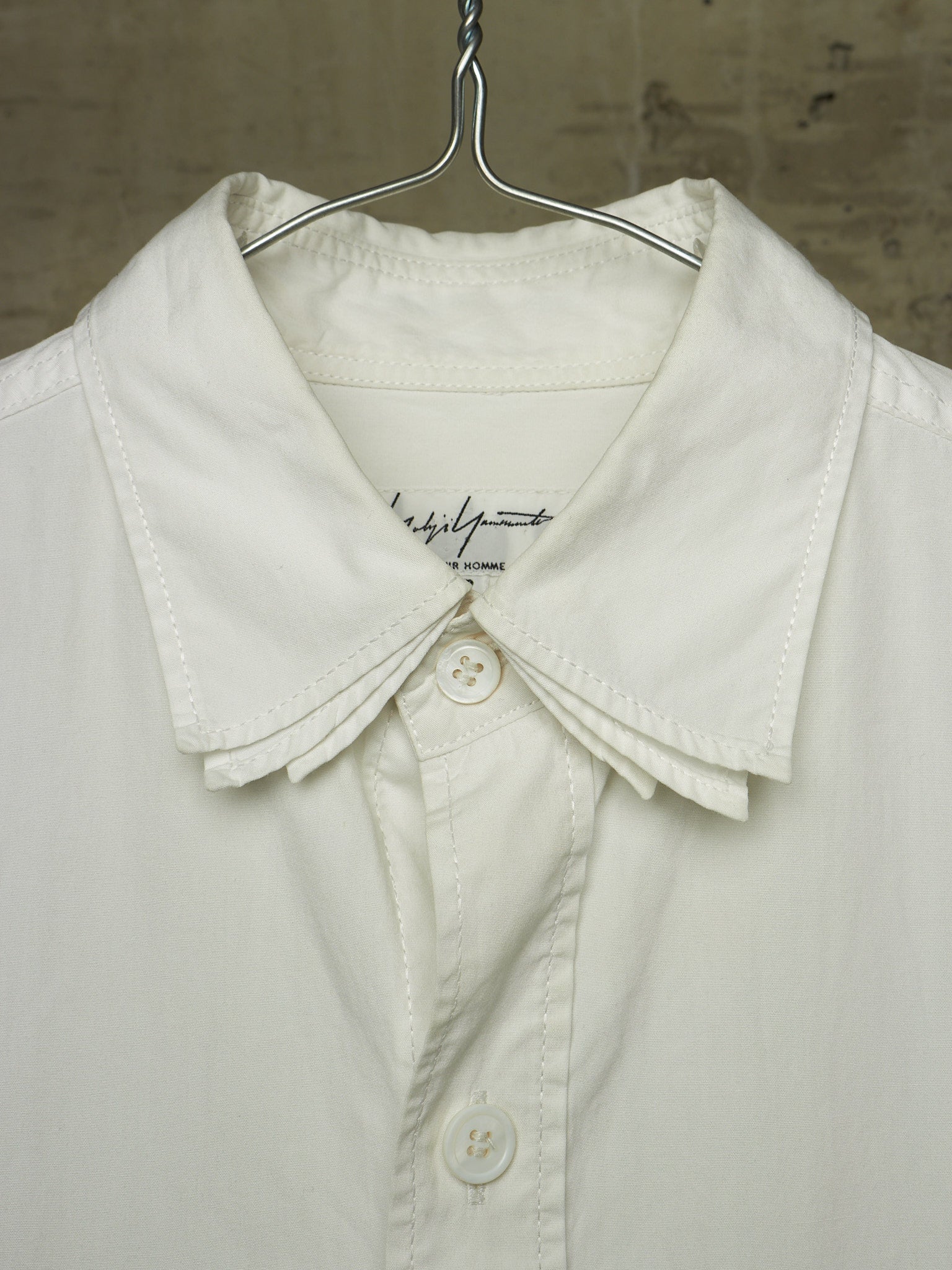 A close-up of a white button-up shirt with a wide, structured collar and decorative pleated detailing at the collar, hanging on a metal hanger against a textured neutral background.