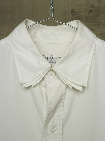 A close-up of a white button-up shirt with a wide, structured collar and decorative pleated detailing at the collar, hanging on a metal hanger against a textured neutral background.