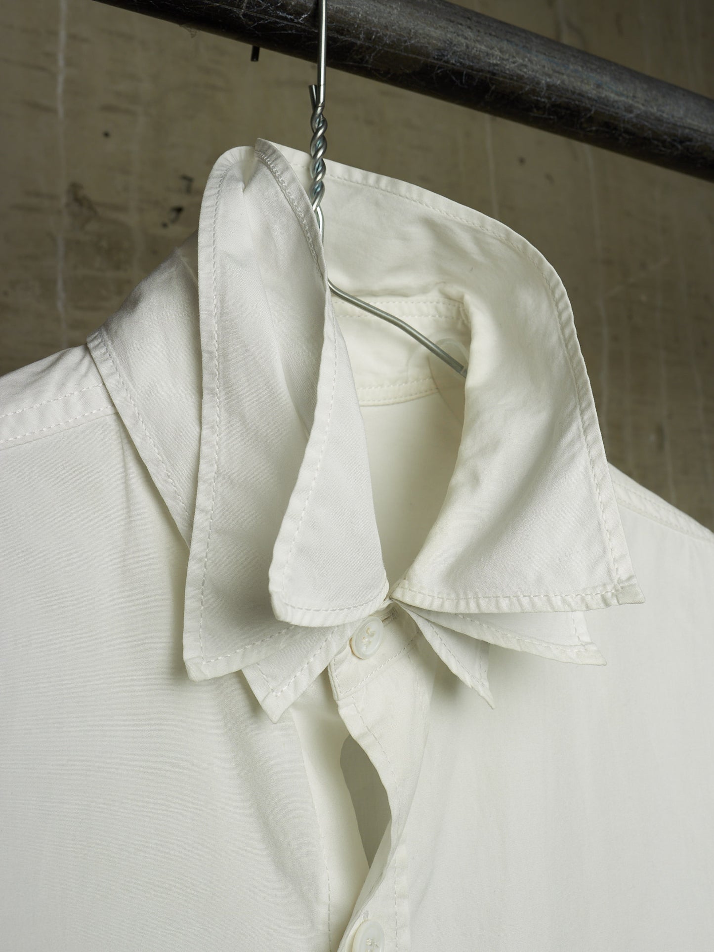 A close-up of a white button-up shirt hanging on a metal hanger from a dark horizontal rod, showing the collar and upper front against a textured beige wall.