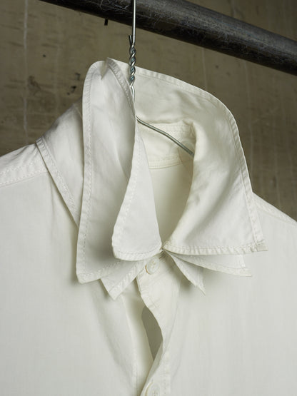 A close-up of a white button-up shirt hanging on a metal hanger from a dark horizontal rod, showing the collar and upper front against a textured beige wall.
