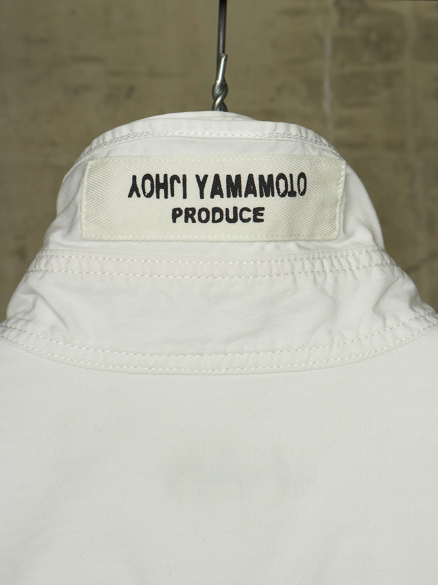 Close-up of the back collar of a white jacket hanging on a metal hanger against a textured beige wall. The collar label reads '֑הושו הרבכך הרול'.