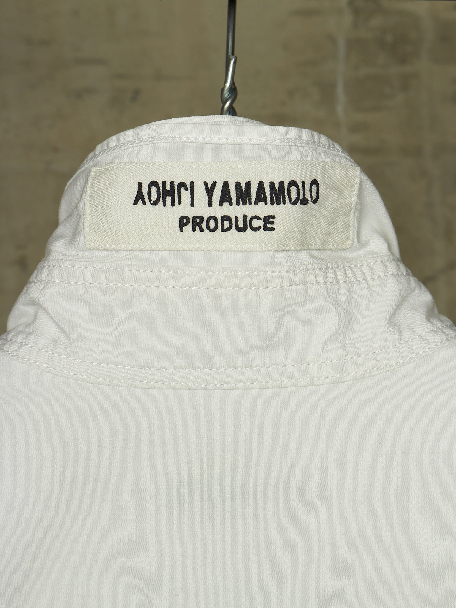 Close-up of the back collar of a white jacket hanging on a metal hanger against a textured beige wall. The collar label reads '֑הושו הרבכך הרול'.