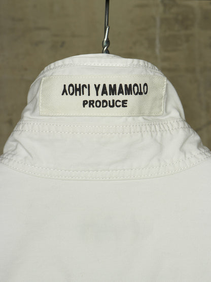 Close-up of the back collar of a white jacket hanging on a metal hanger against a textured beige wall. The collar label reads '֑הושו הרבכך הרול'.