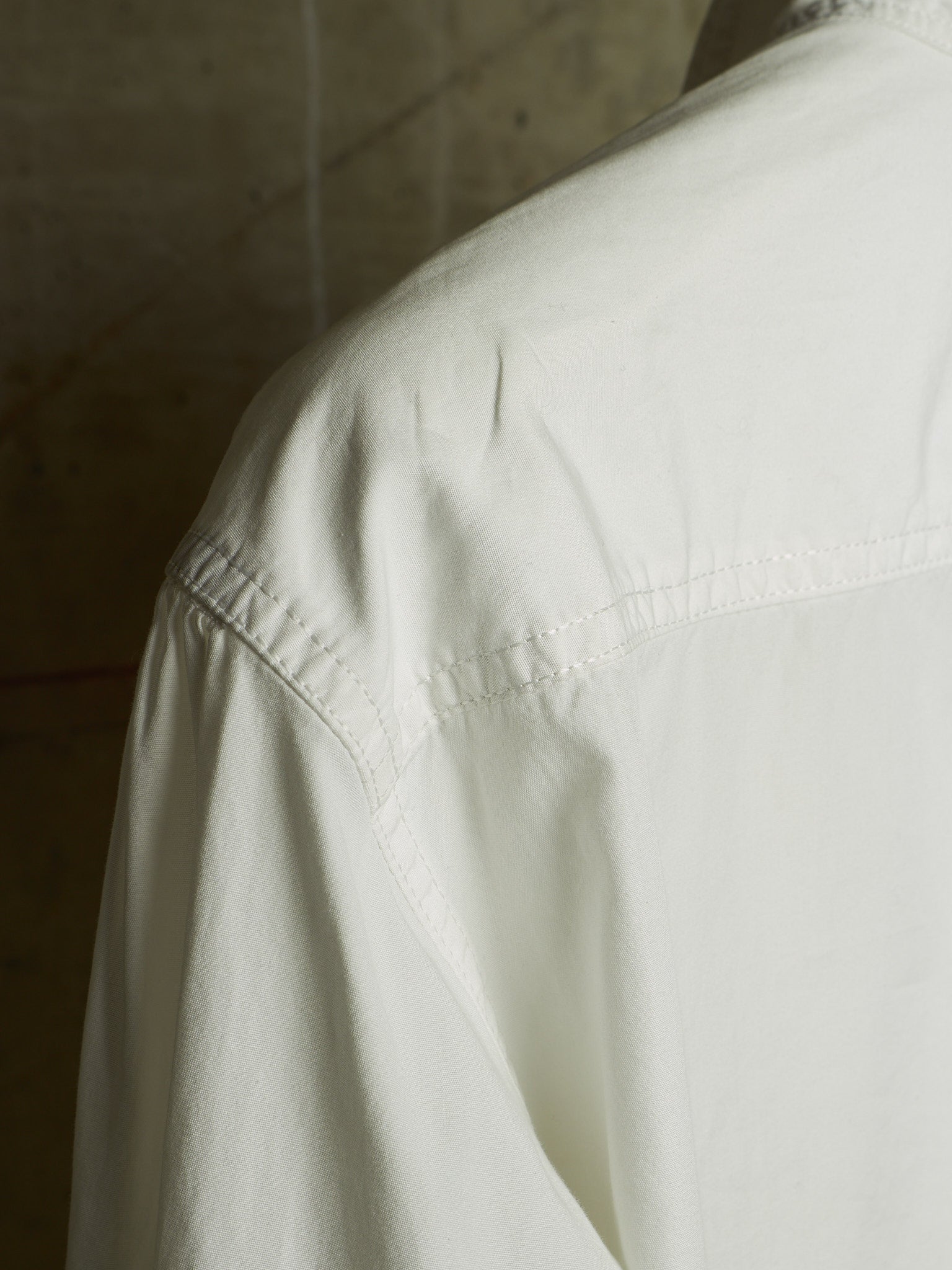 Close-up of a white cotton shirt showing the shoulder seam, sleeve, and chest stitching against a textured concrete background.