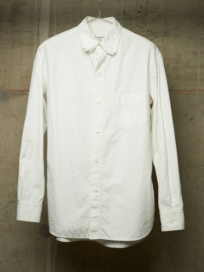 An off-white long-sleeve button-up shirt with a chest pocket, hanging on a metal hanger against a textured concrete wall.
