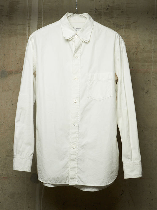 An off-white long-sleeve button-up shirt with a chest pocket, hanging on a metal hanger against a textured concrete wall.