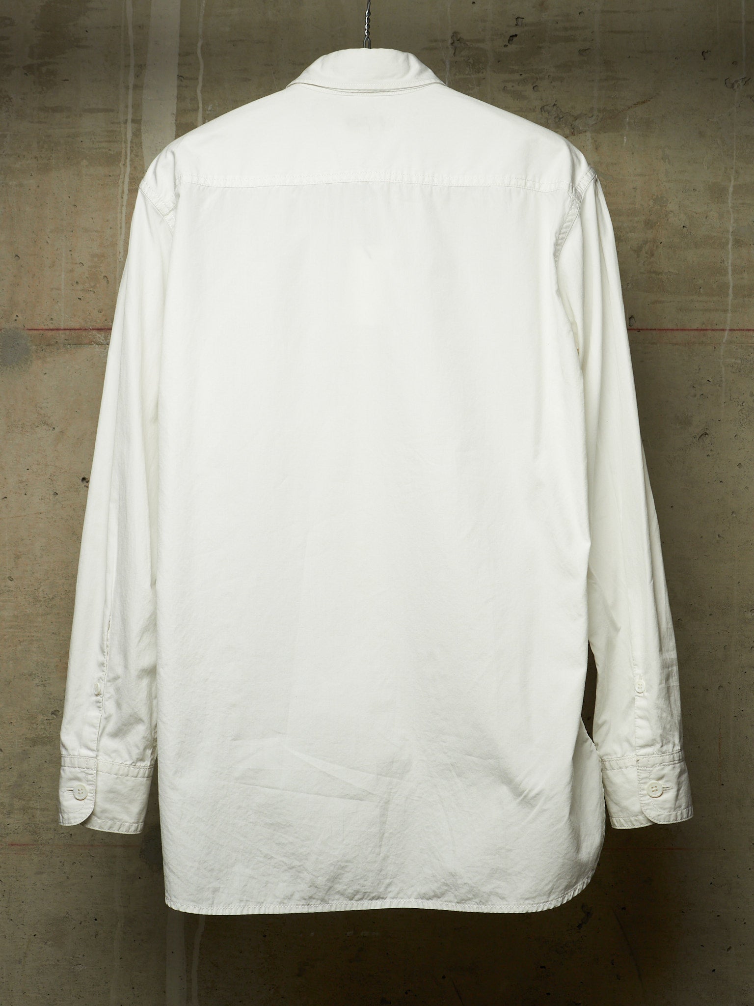 A white long-sleeve button-up shirt shown from the back, hanging against a textured concrete wall.