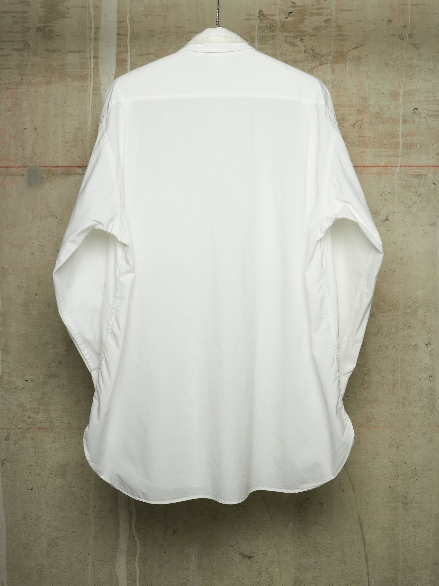 A white oversized long-sleeve shirt shown from the back, hanging against a textured concrete wall.