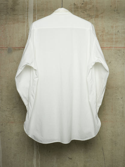 A white oversized long-sleeve shirt shown from the back, hanging against a textured concrete wall.