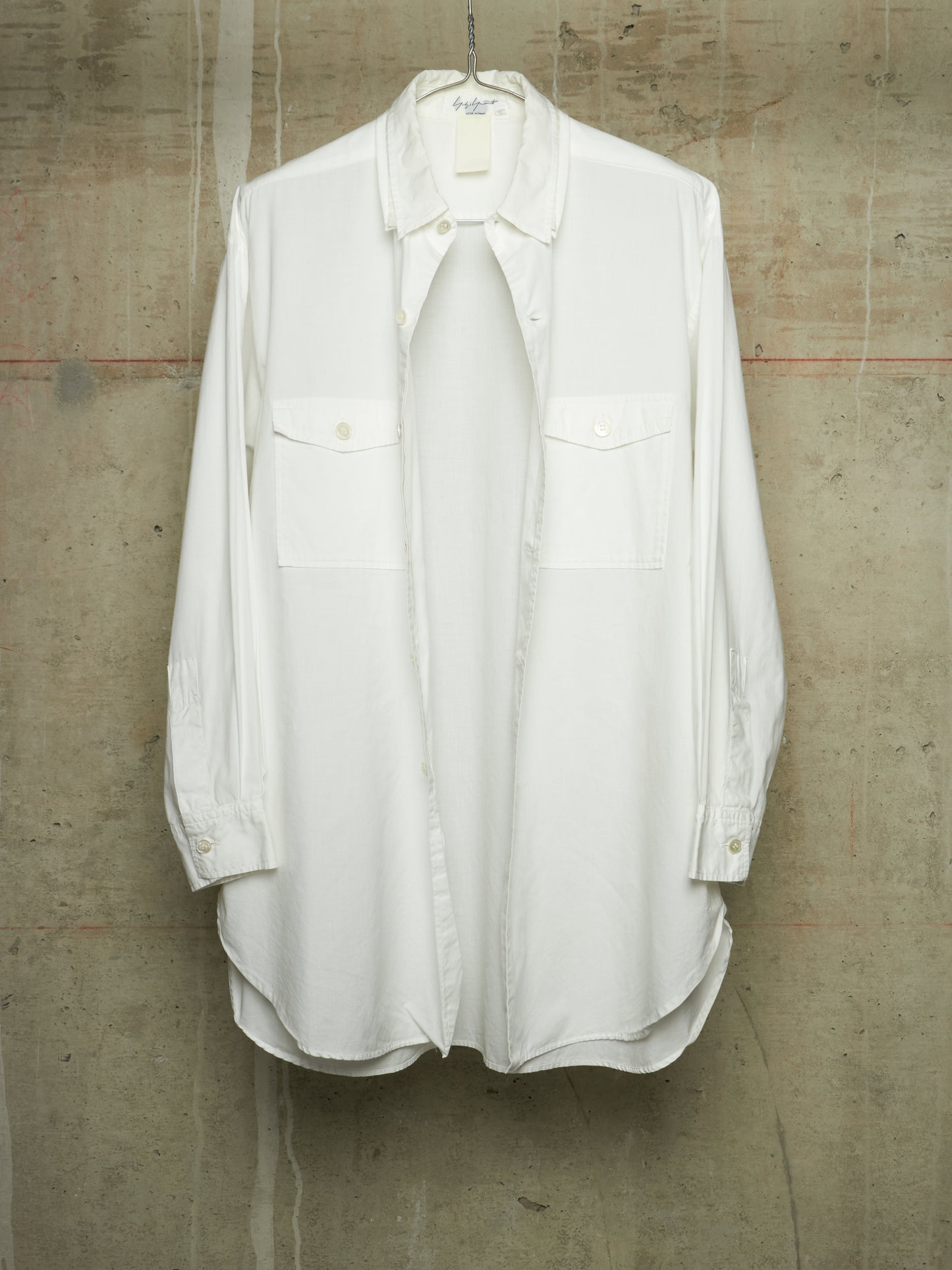 A white long-sleeve button-up shirt with two chest flap pockets, hanging on a metal hanger against a textured concrete wall.