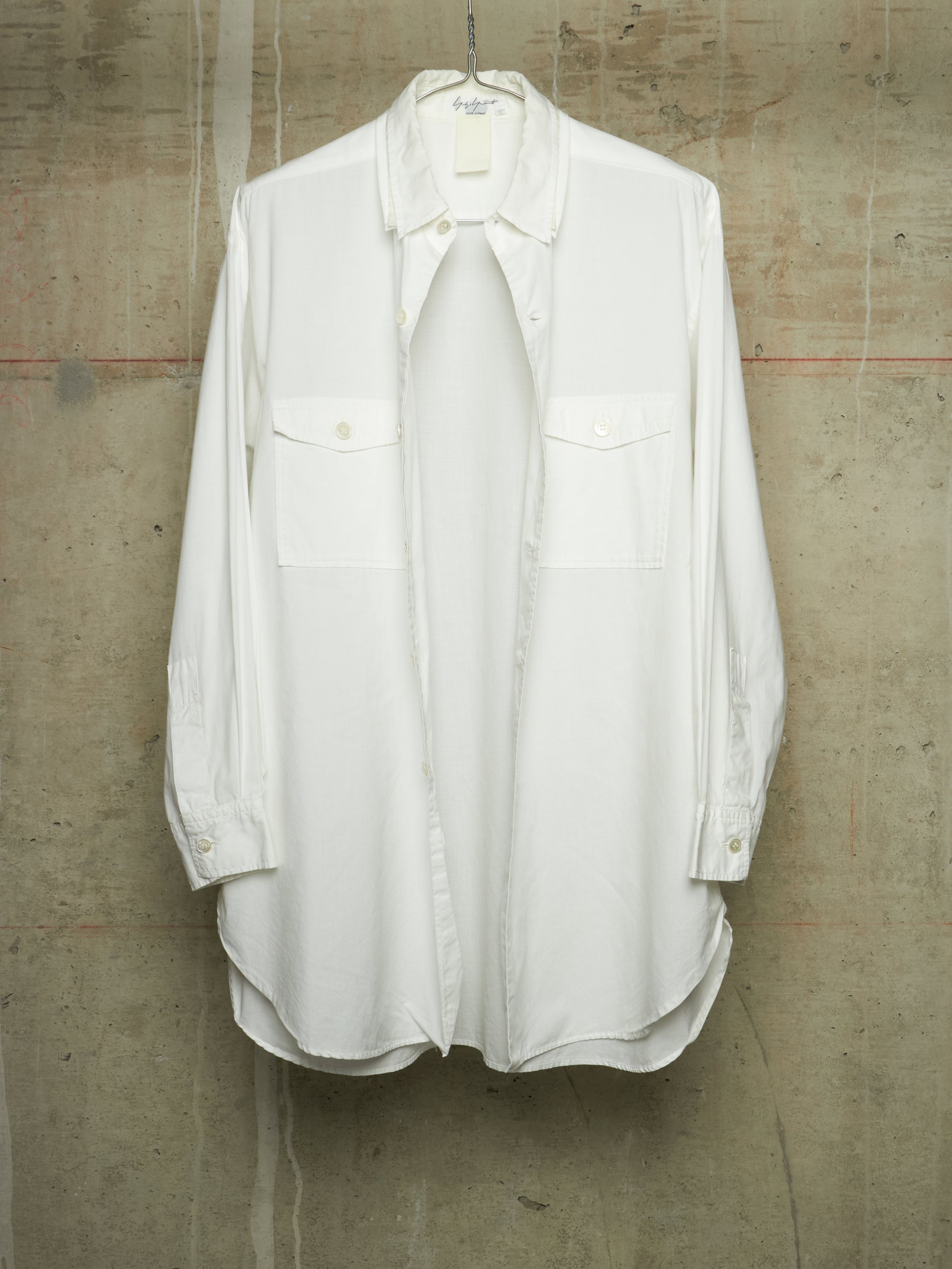 A white long-sleeve button-up shirt with two chest flap pockets, hanging on a metal hanger against a textured concrete wall.