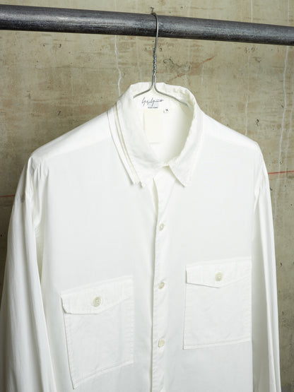 A white long-sleeve button-up shirt with two chest flap pockets, hanging on a metal hanger from a dark horizontal rod against a textured concrete wall.