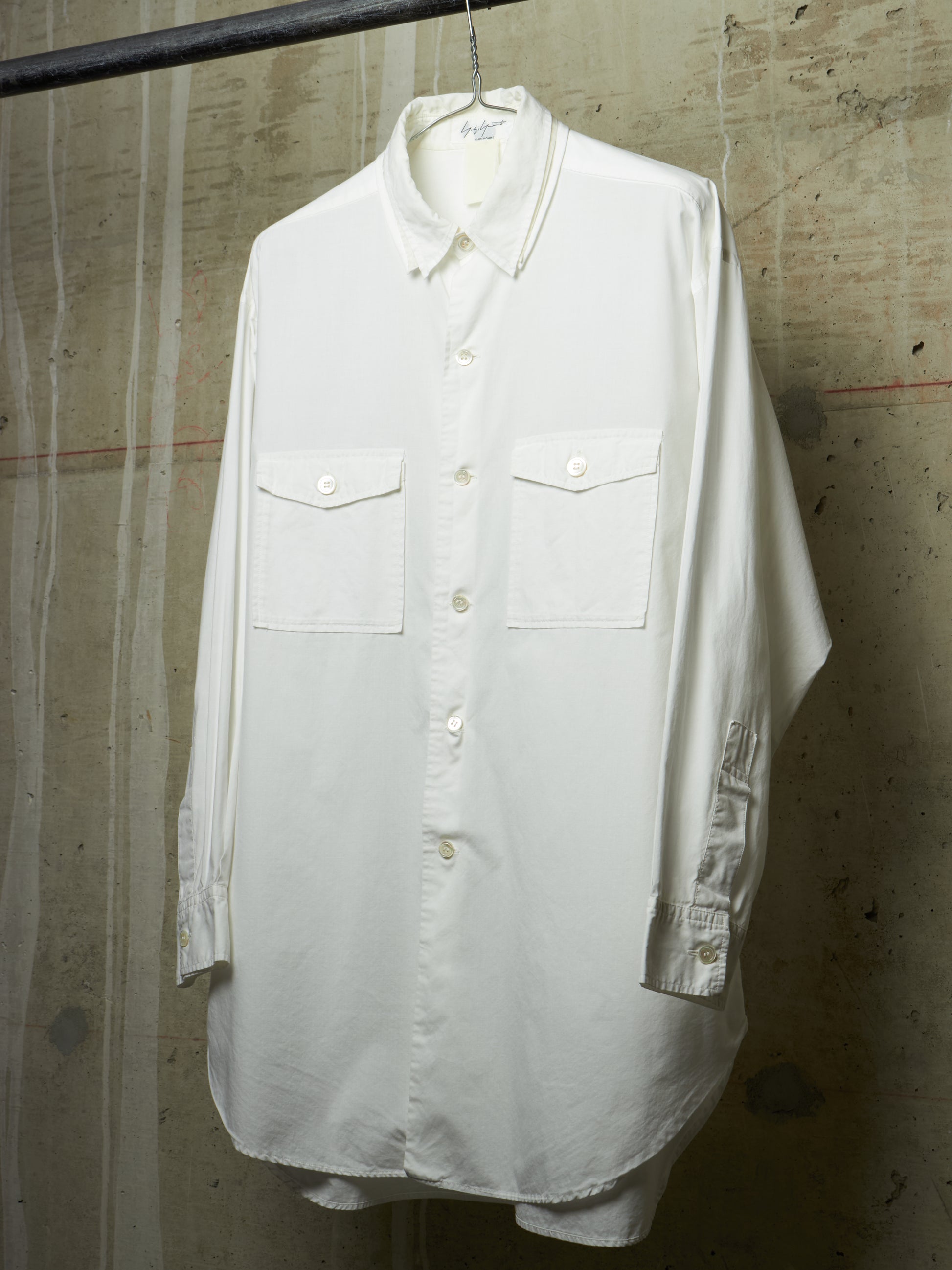 A white long-sleeve button-up shirt with two chest flap pockets, hanging on a hanger against a textured concrete wall.