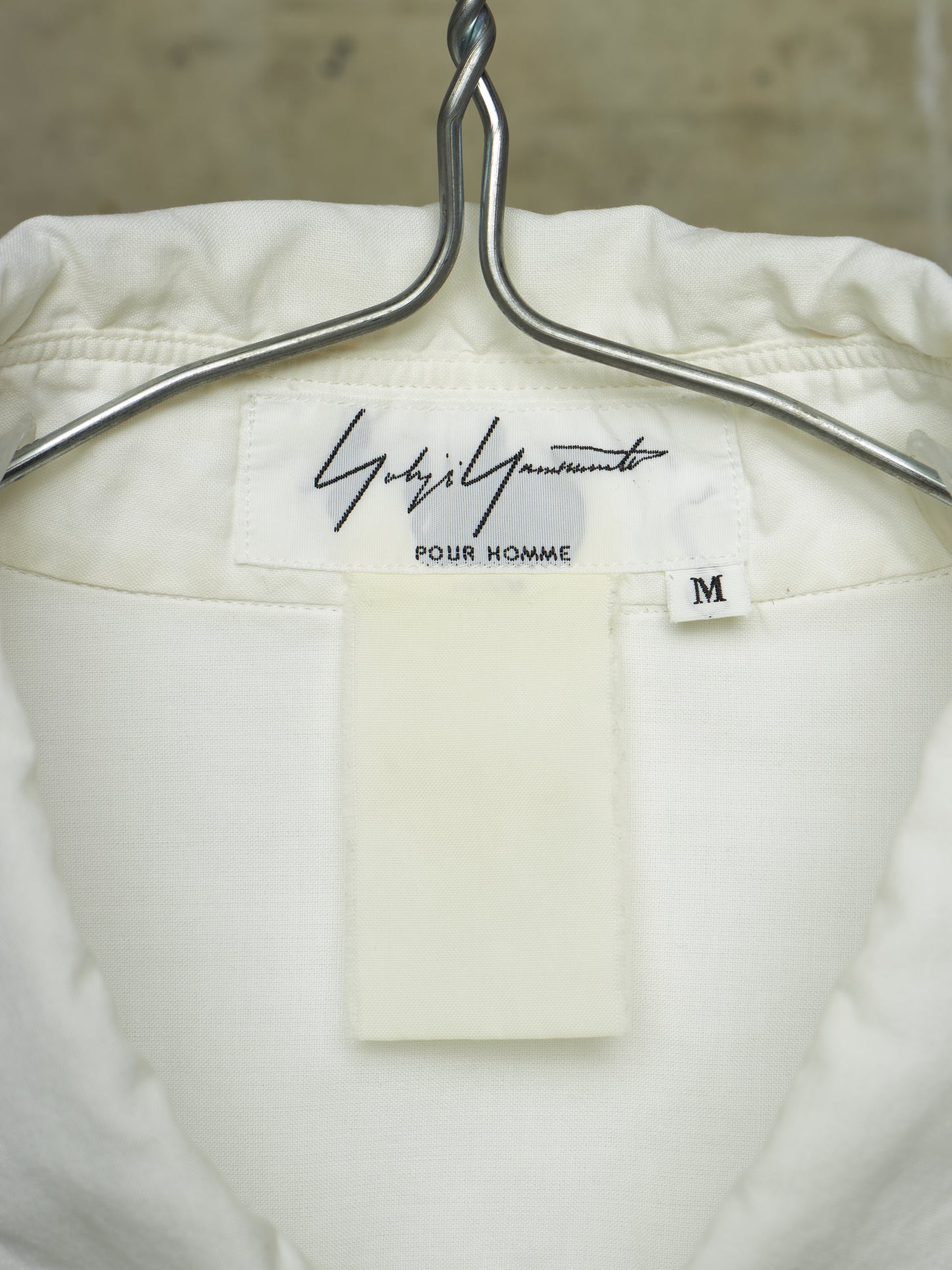 Close-up of the inside neck of a white or off-white jacket hanging on a silver metal hanger. The inner label reads 'Gucci' in script with 'Pour Homme' underneath and a small size tag marked 'M'.