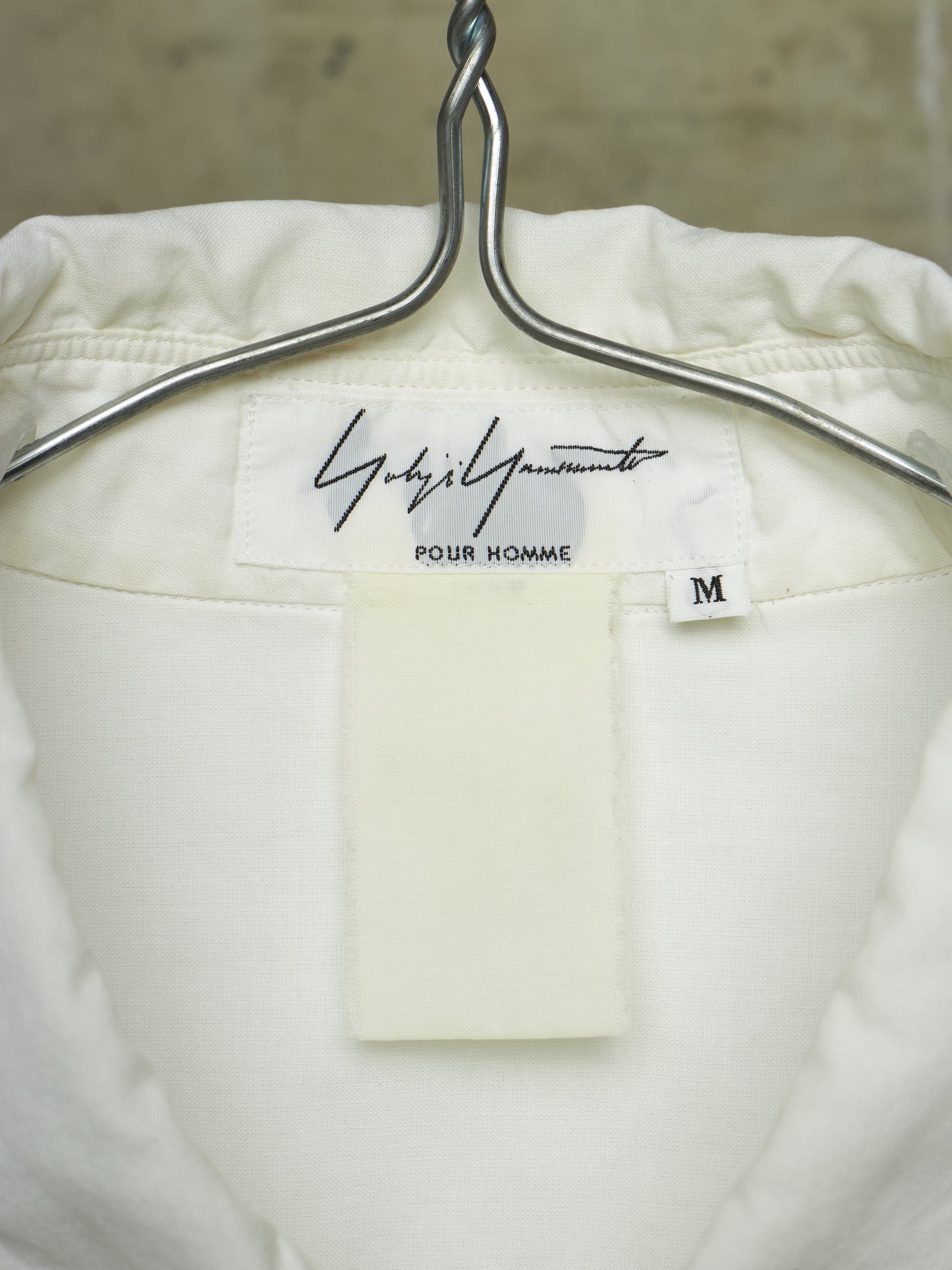 Close-up of the inside neck of a white or off-white jacket hanging on a silver metal hanger. The inner label reads 'Gucci' in script with 'Pour Homme' underneath and a small size tag marked 'M'.
