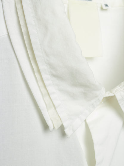 Close-up detail of a white or off-white fabric garment, showing layered pleats and a small cream hangtag attached near the collar area.