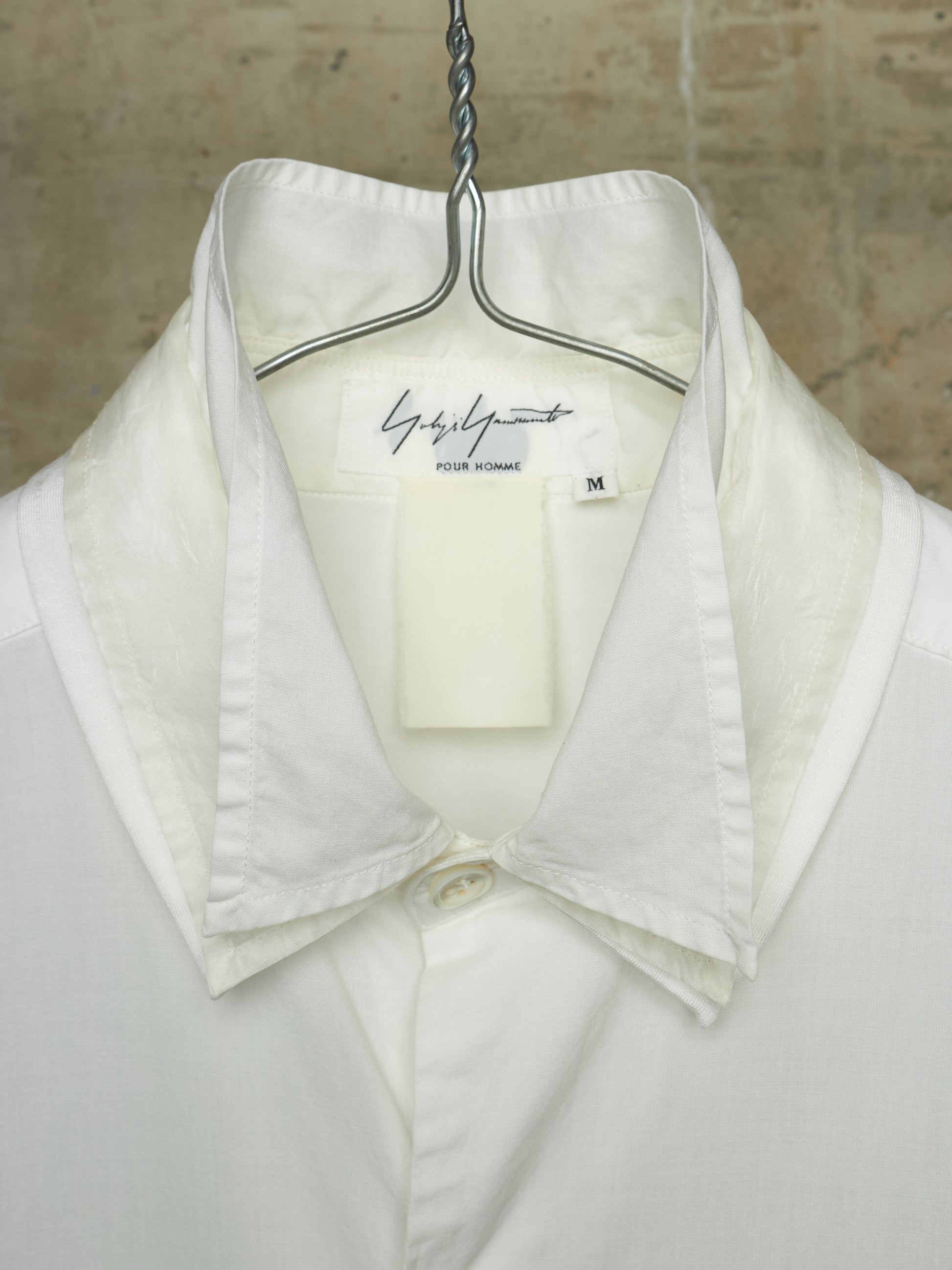 A white collared button-up shirt hanging on a metal hanger against a textured beige wall. The inner neck label shows the brand 'Gisèle Paris' and a size 'M'.