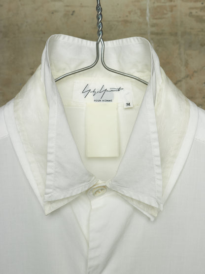 A white collared button-up shirt hanging on a metal hanger against a textured beige wall. The inner neck label shows the brand 'Gisèle Paris' and a size 'M'.