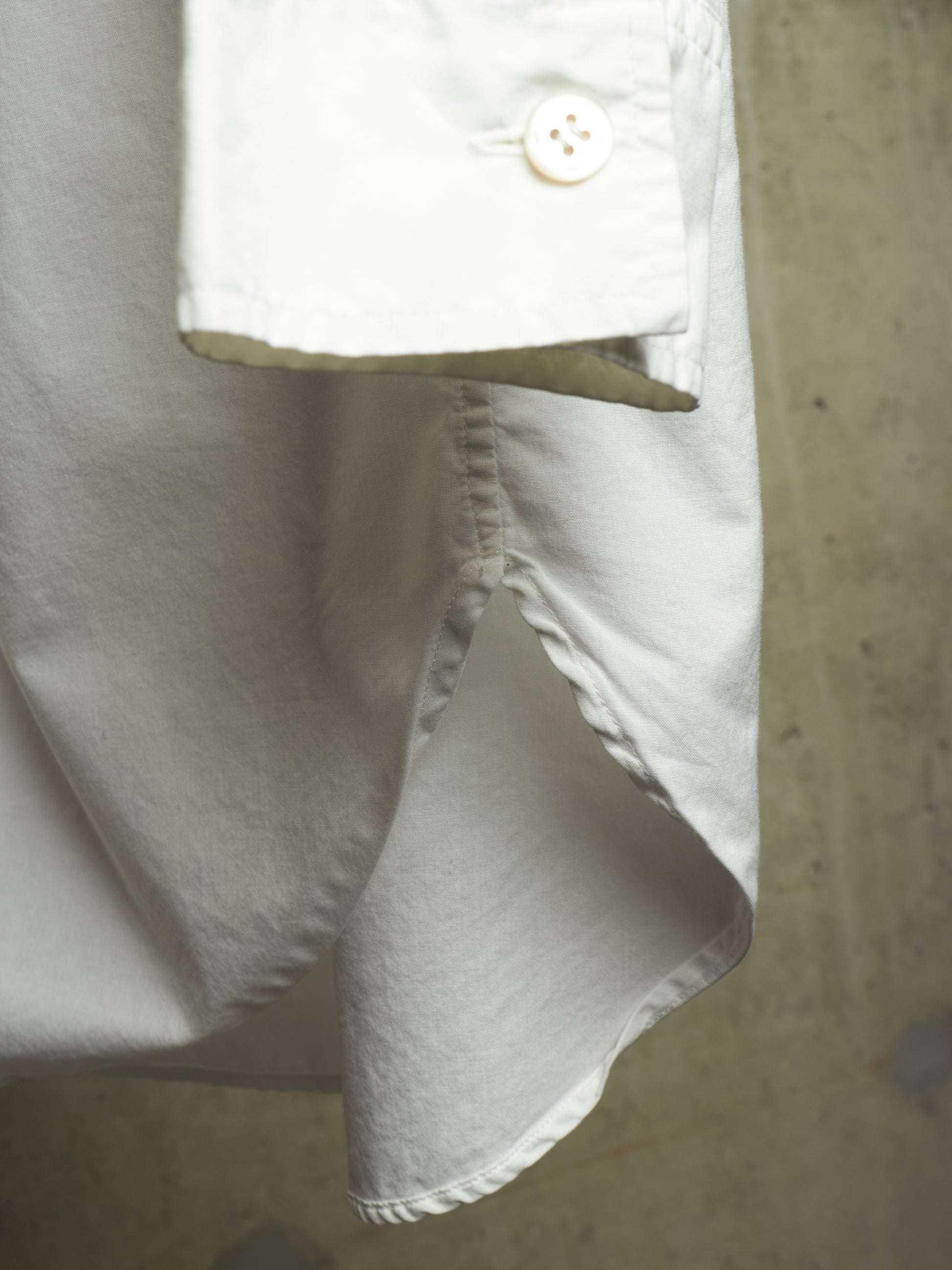 Close-up detail of a white shirt or blouse showing the underside of a buttoned cuff, a raw hem, and stitched fold, photographed against a neutral concrete-like background.