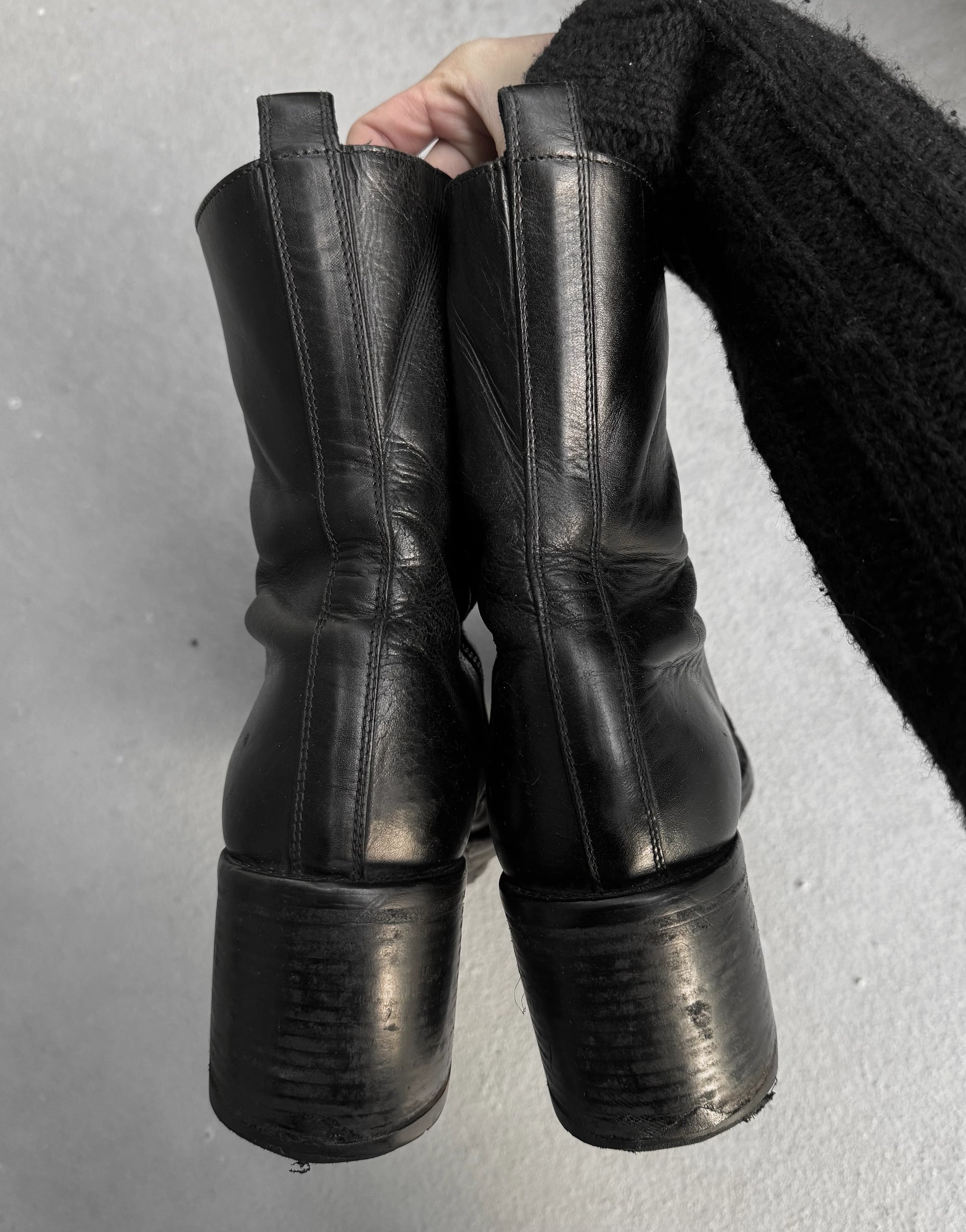 Black leather knee-high boots held by a person against a gray background