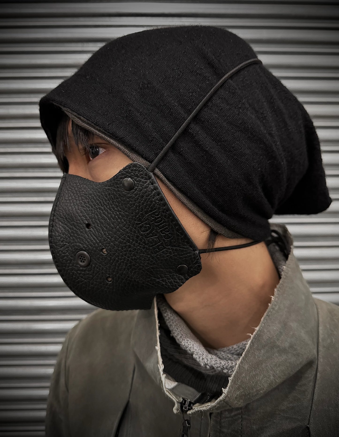 Person wearing a black leather face mask and hood against a metallic background