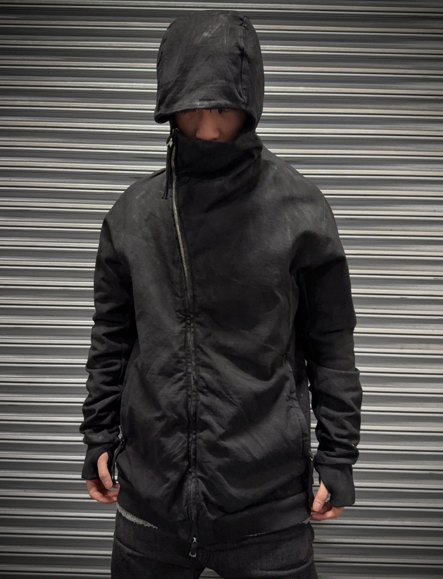 Person wearing a black hooded jacket in front of a metallic shutter.