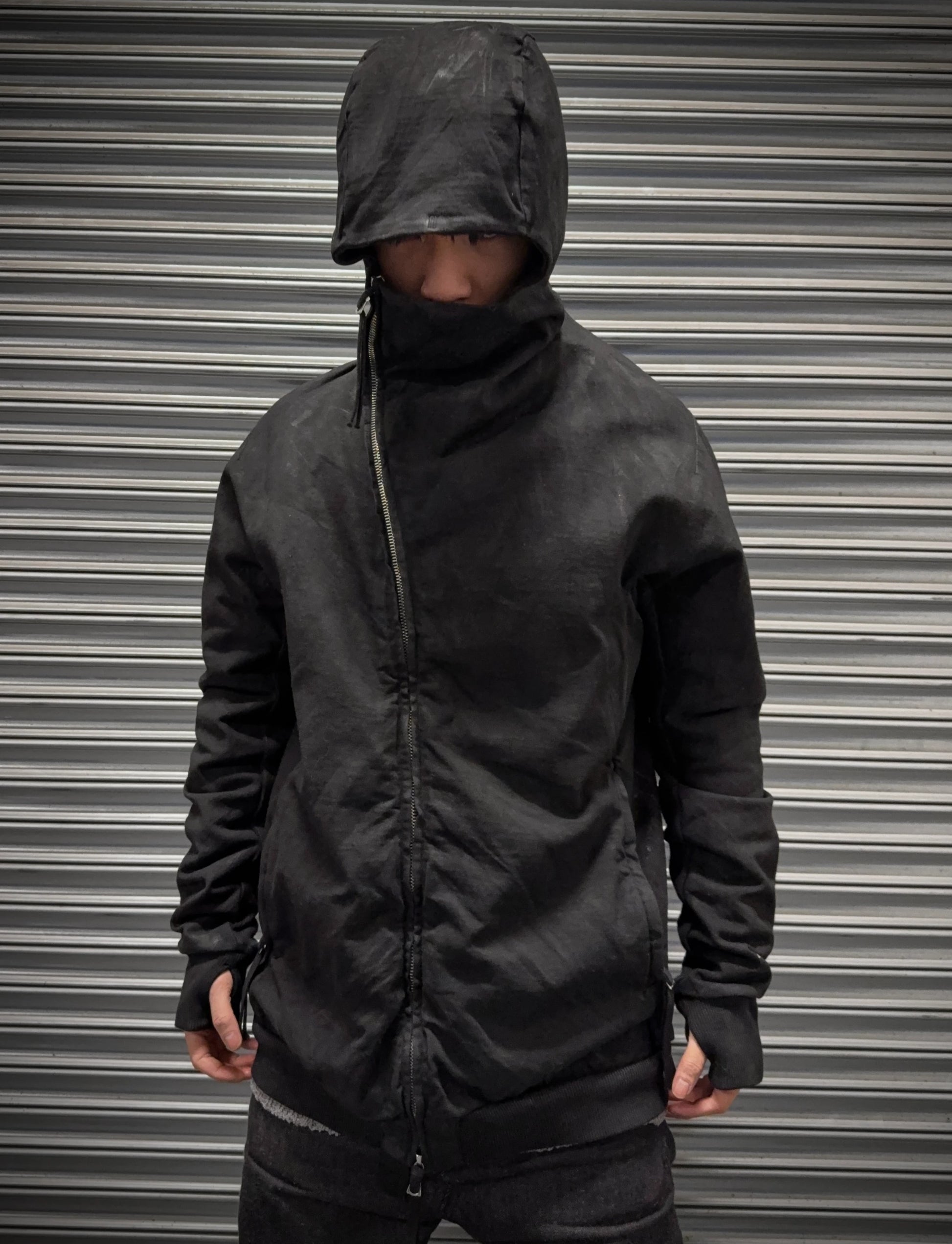 Person wearing a black hooded jacket in front of a metallic shutter.