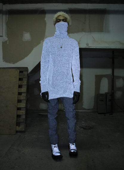 Person wearing a light gray sweater, dark pants, and black gloves in a dimly lit room.