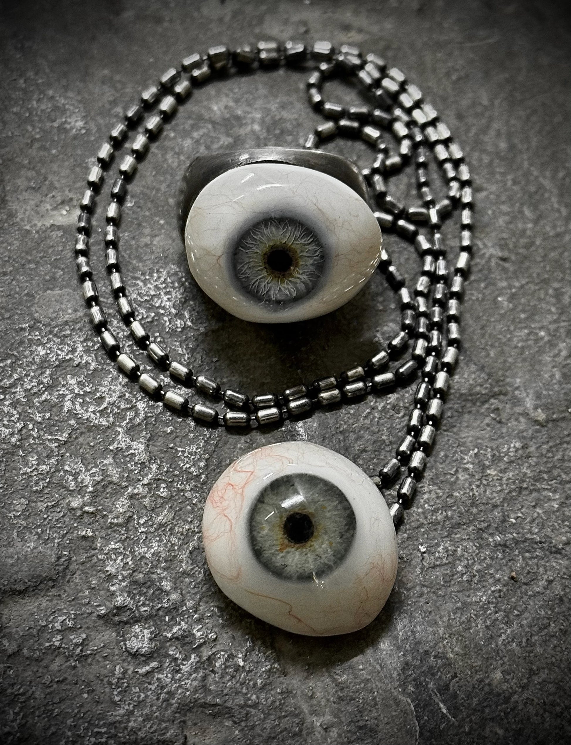Two eyeball-shaped pendants on a chain against a textured gray background