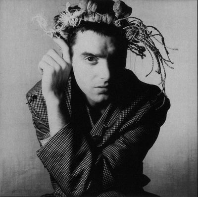 Black and white portrait of British fashion designer Christopher Nemeth standing against a grey background with a crown of fl