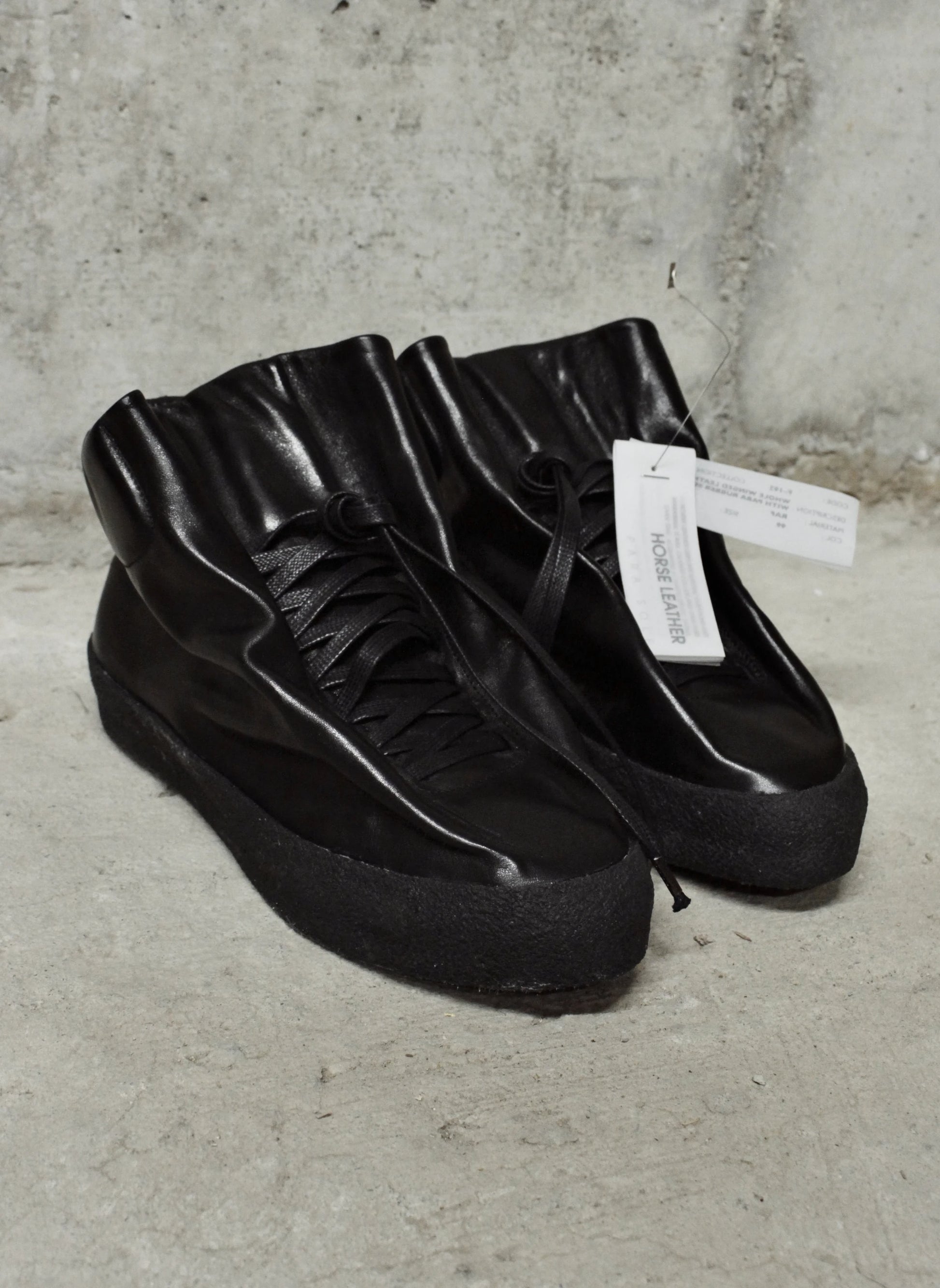 Pair of black high-top sneakers on a concrete floor with a visible brand tag.