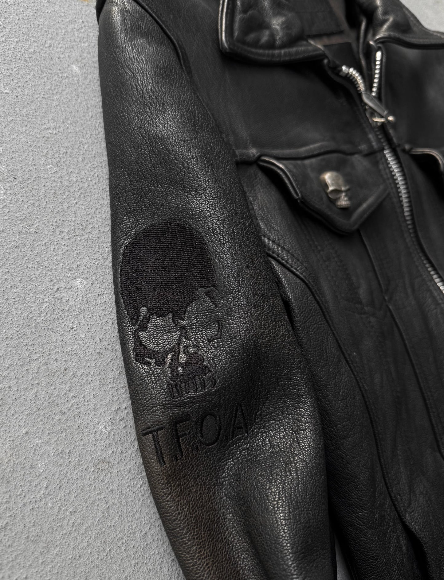 Black leather jacket with skull design and 'T.F.O.A.' branding on a gray background