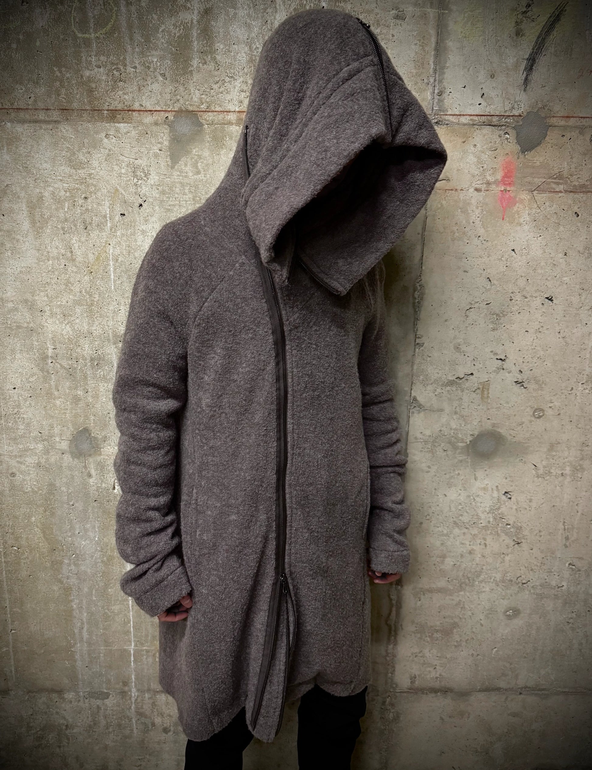 Person wearing a long, dark gray hooded coat against a textured wall.